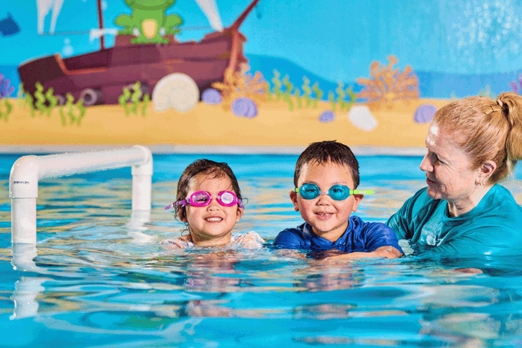 A woman with blonde hair is standing in a swimming pool in water is up to her shoulders. She's a swim instructor next to two children in the water with her. The children are preschool age, one is wearing pink goggles and one is wearing blue goggles. Everyone is smiling and having fun.