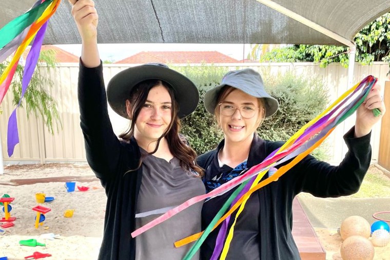 Two smiling early childhood educators holding colorful ribbons in an outdoor play area